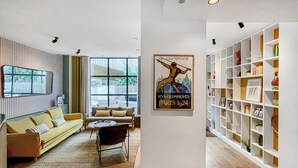 Un salón moderno con sofá, sillón y mesa de centro. Se ve un ventanal con vistas a la calle. También se aprecia un cartel en la pared y una estantería con diversos objetos.