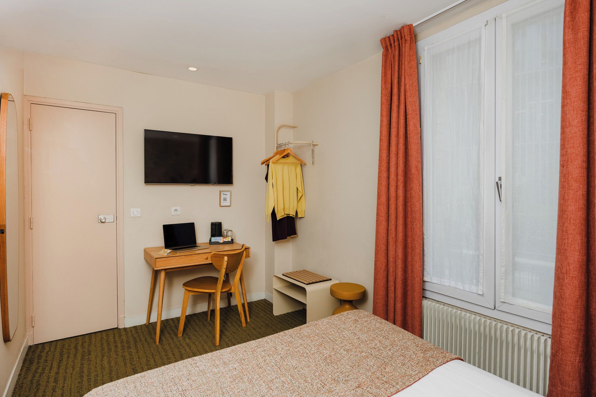 Une chambre d’hôtel comprenant un lit, un bureau avec un ordinateur portable, une chaise, un téléviseur fixé au mur, une fenêtre avec des rideaux et un radiateur.