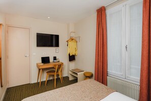 Une chambre d’hôtel comprenant un lit, un bureau avec un ordinateur portable, une chaise, un téléviseur fixé au mur, une fenêtre avec des rideaux et un radiateur.