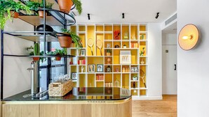 Un área de recepción moderna con un mostrador de madera, una planta en maceta, una vitrina de vidrio y una estantería amarilla y blanca con diversos objetos.
