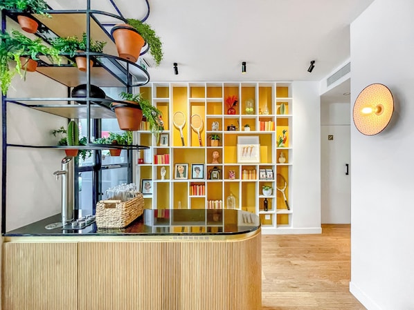 Un área de recepción moderna con un mostrador de madera, una planta en maceta, una vitrina de vidrio y una estantería amarilla y blanca con diversos objetos.