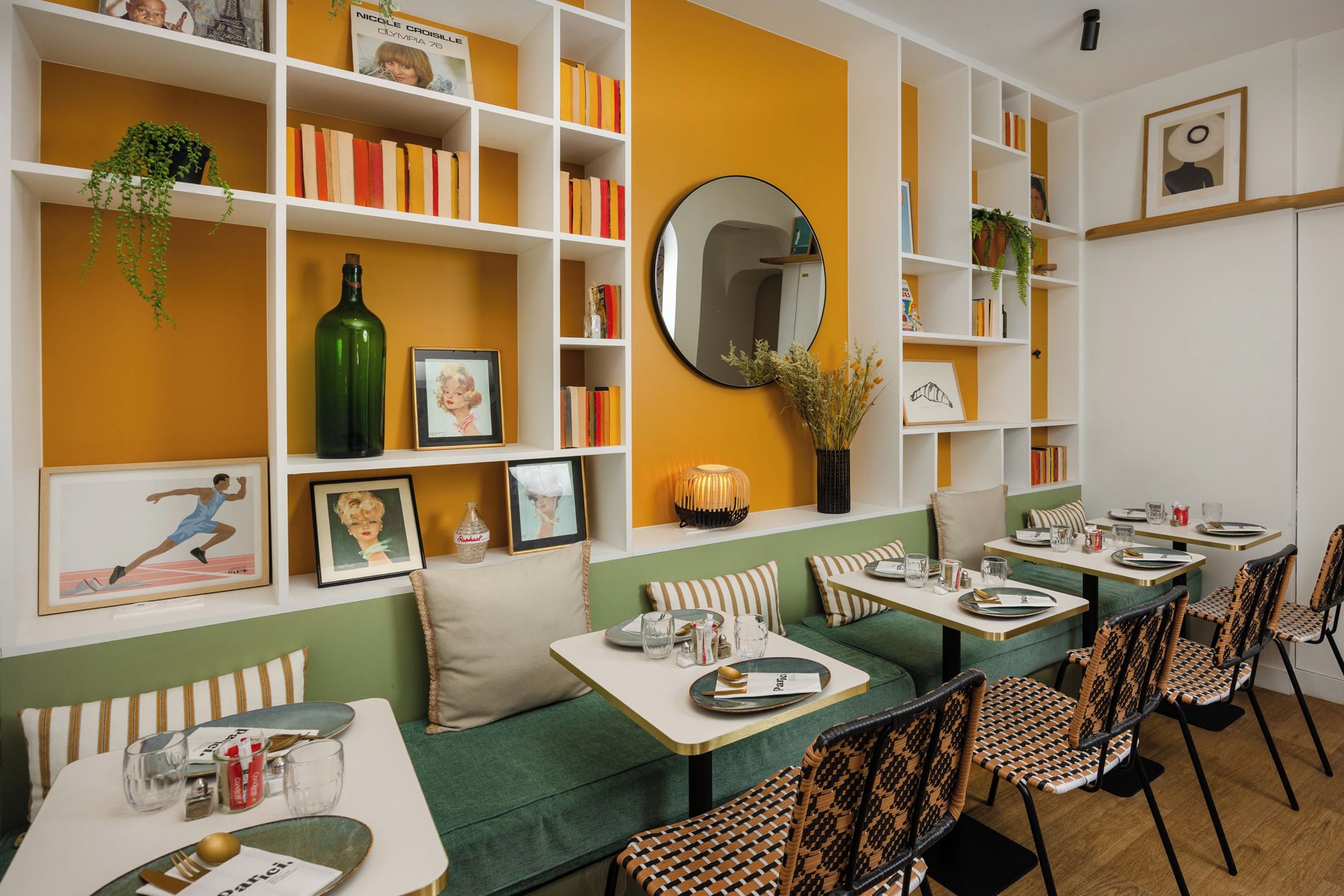 Un coin repas avec des tables préparées pour deux personnes, une bibliothèque contenant divers objets et un miroir accroché au mur.