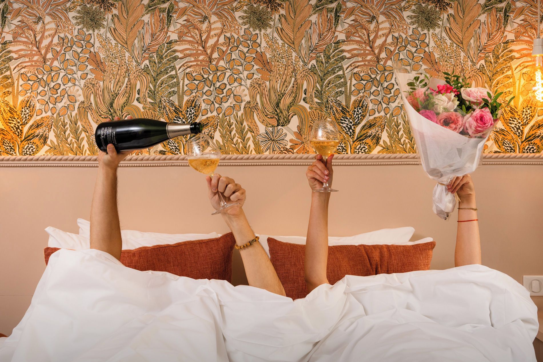 Une personne allongée dans son lit, une bouteille de champagne et un verre de champagne à portée de main, ainsi qu'un bouquet de fleurs.