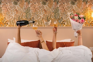 Une personne allongée dans son lit, une bouteille de champagne et un verre de champagne à portée de main, ainsi qu'un bouquet de fleurs.