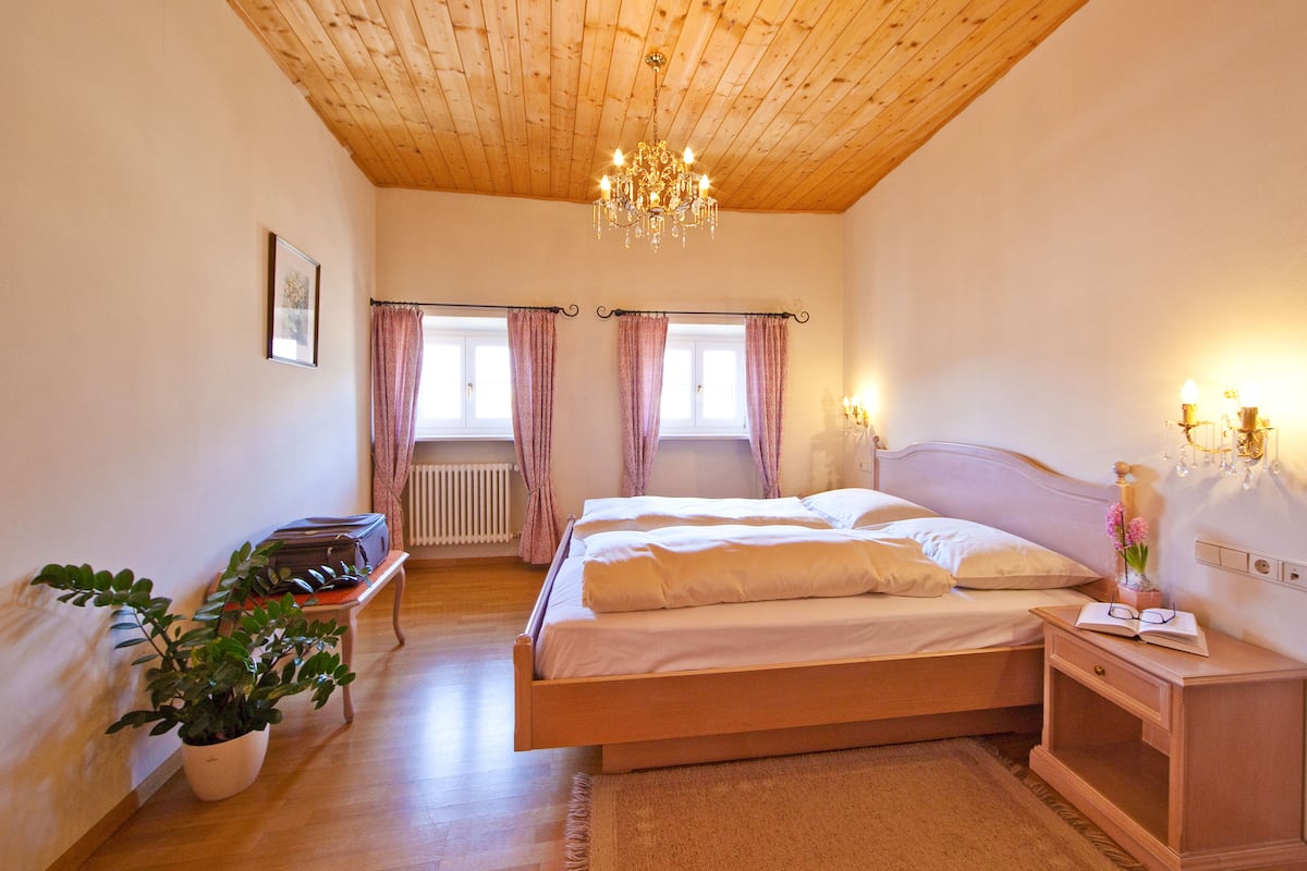 Un dormitorio con techo de madera, una cama con ropa blanca, una mesita de noche con una lámpara y una planta en maceta.