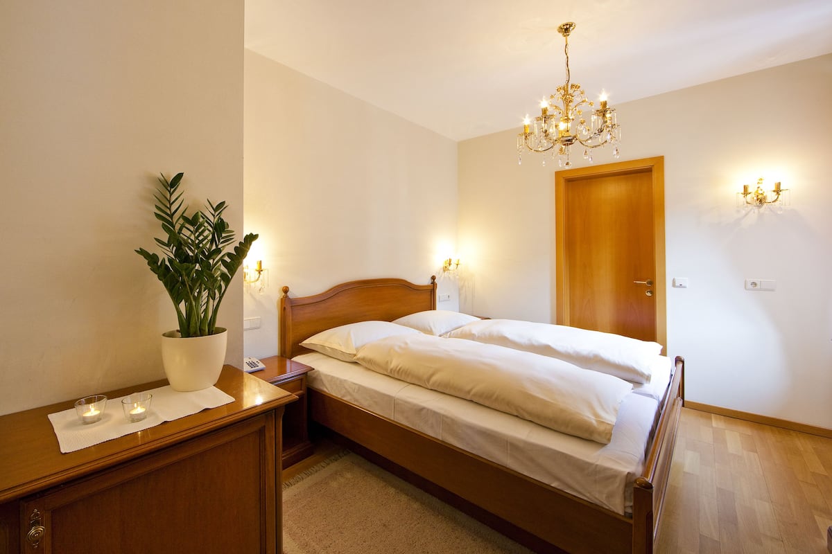 Una cama bien hecha en una habitación con cabecera de madera, mesita de noche con una planta, candelabro y apliques de pared.