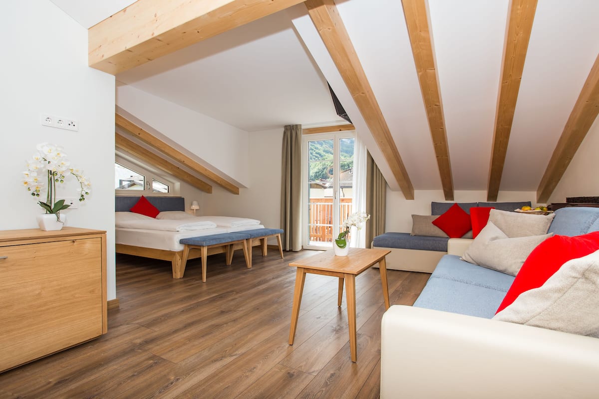 Una habitación moderna estilo loft con techo de madera, un sofá, una cama, una mesita y vistas al exterior con vegetación.