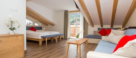 Ein modernes Loft-Zimmer mit Holzboden, einer Couch, einem Bett, einem kleinen Tisch und Blick auf Grünflächen.