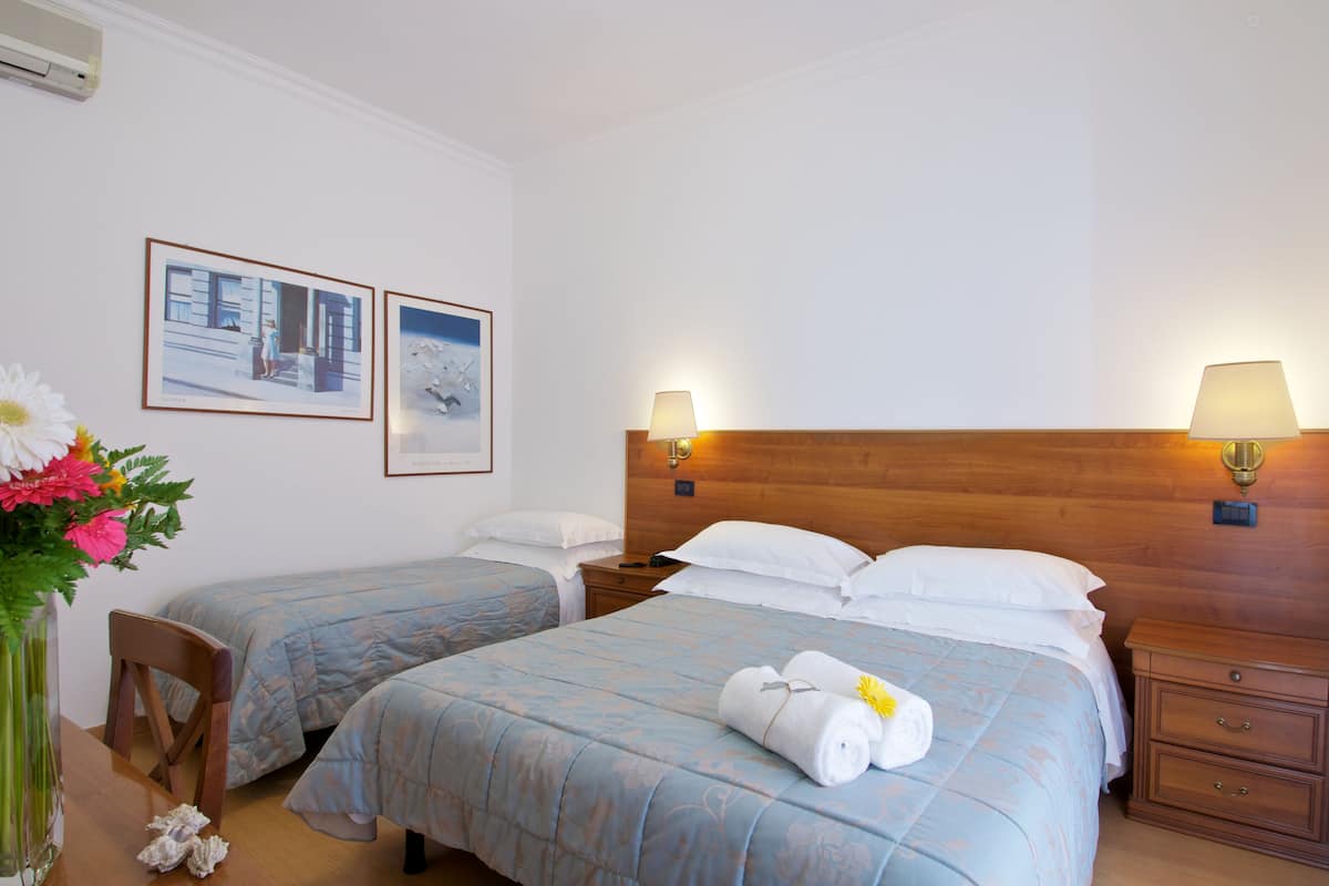 A hotel room with two beds, a wooden headboard, bedside lamps, a nightstand, a vase with flowers, and framed pictures on the wall.