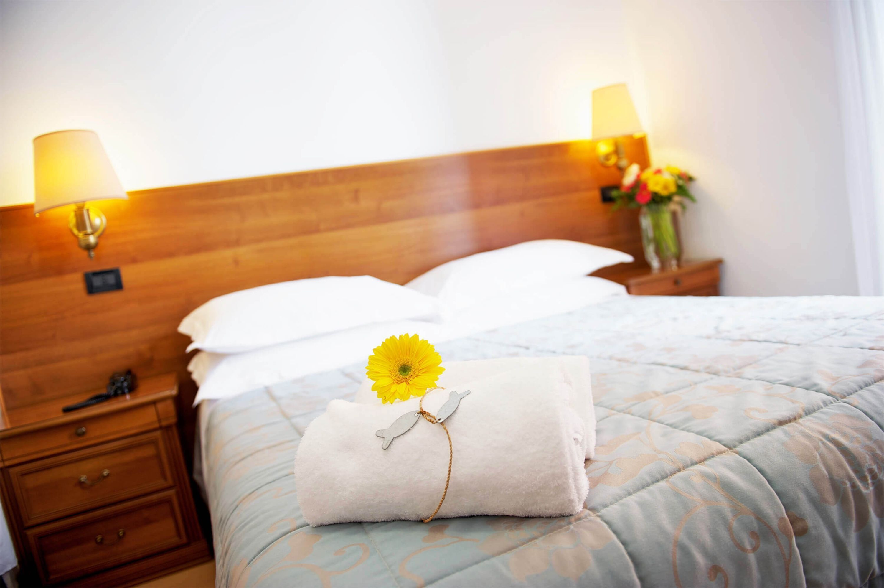 A neatly made bed with a folded towel and a yellow flower, two bedside lamps, and a wooden headboard.