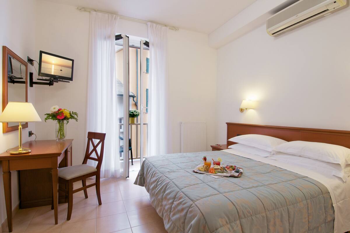 A hotel room with a bed, a desk with a lamp, a chair, a TV, a window with curtains, and a wall-mounted air conditioner.