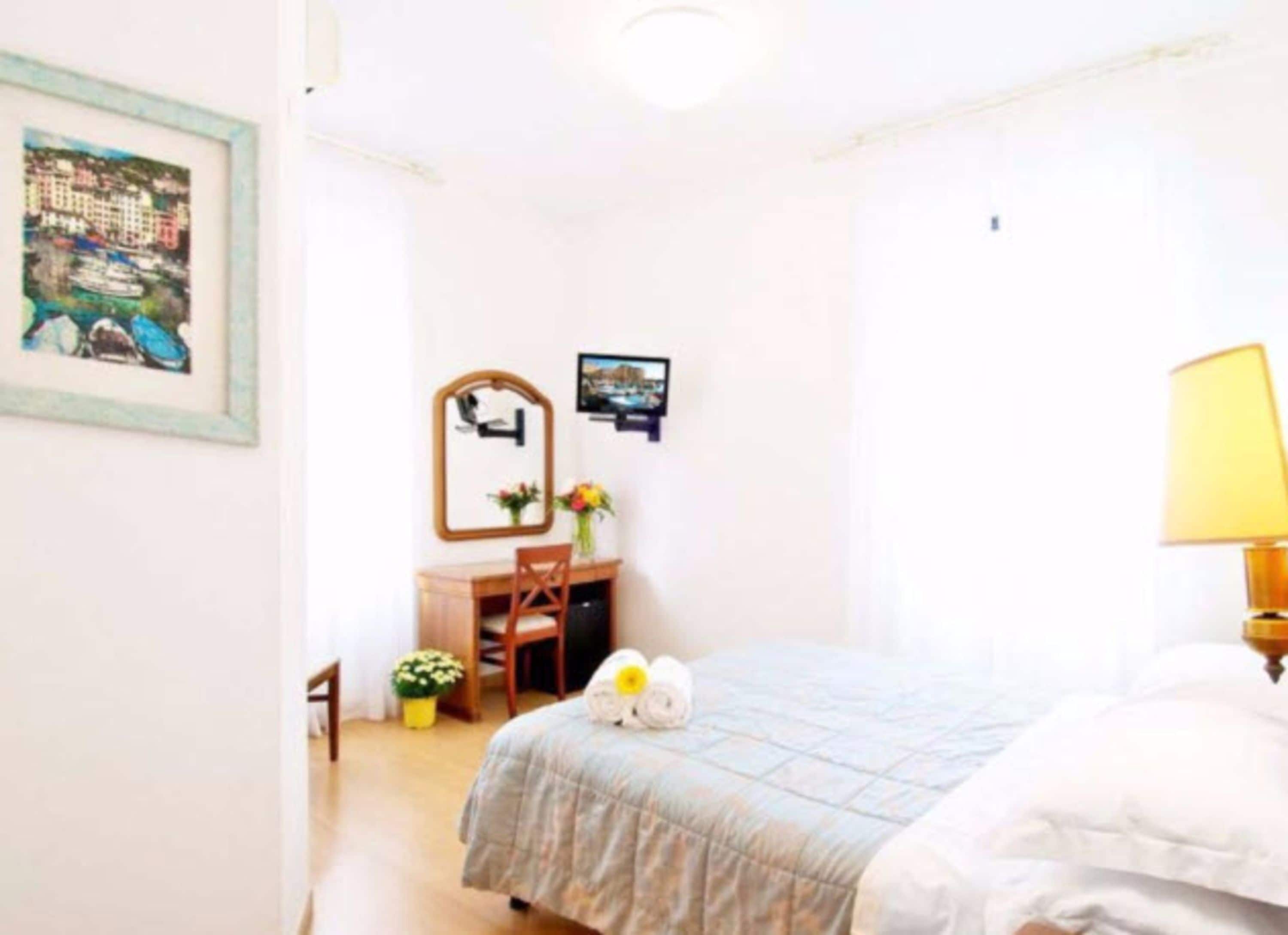 A hotel room with a bed, a desk, a chair, a lamp, and a framed picture on the wall.