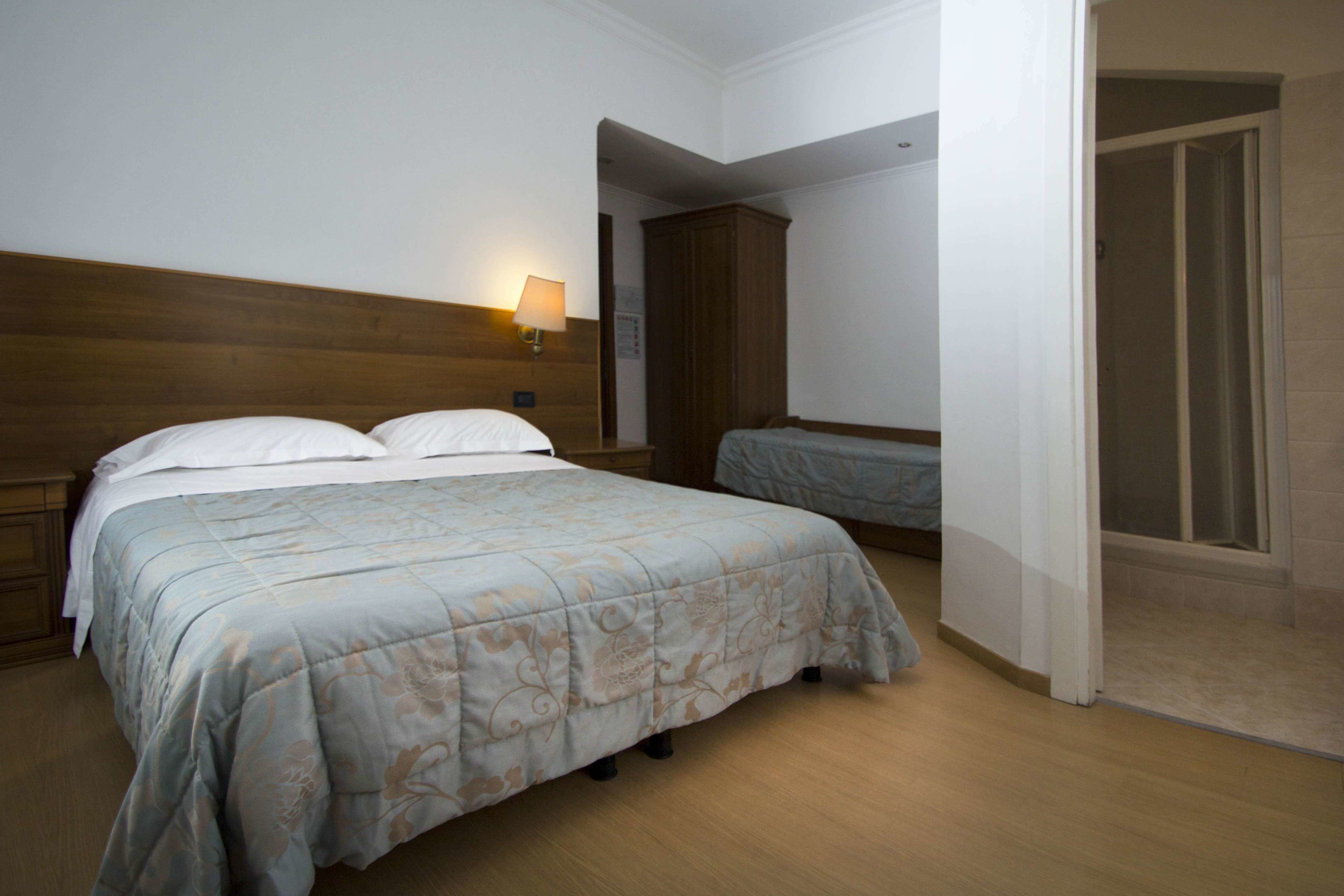 Un dormitorio con cabecera de madera, cama con colcha estampada, mesita de noche con lámpara y una puerta que da a otra habitación.