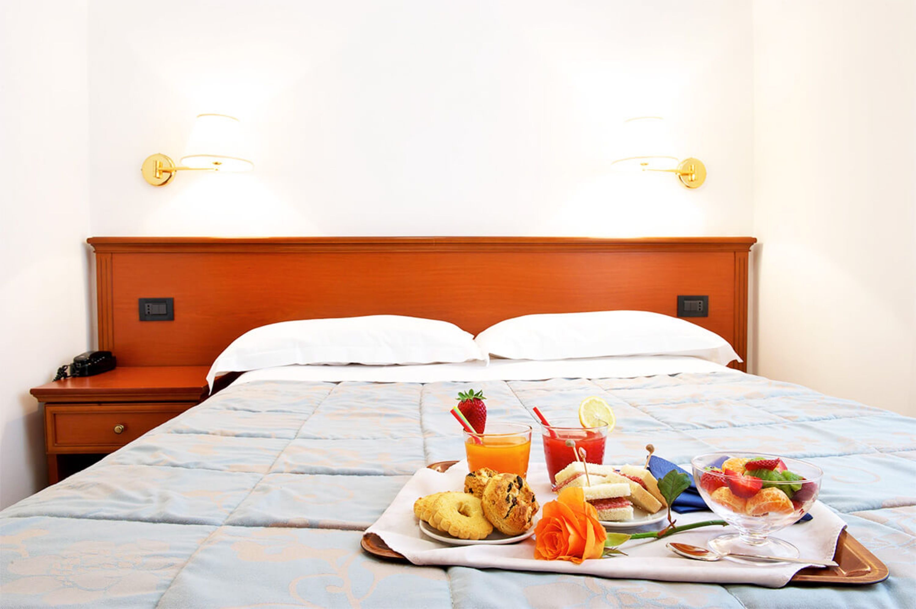 A hotel room with a neatly made bed, a bedside table with a phone, and a tray with breakfast items including pastries, fruit, and drinks.