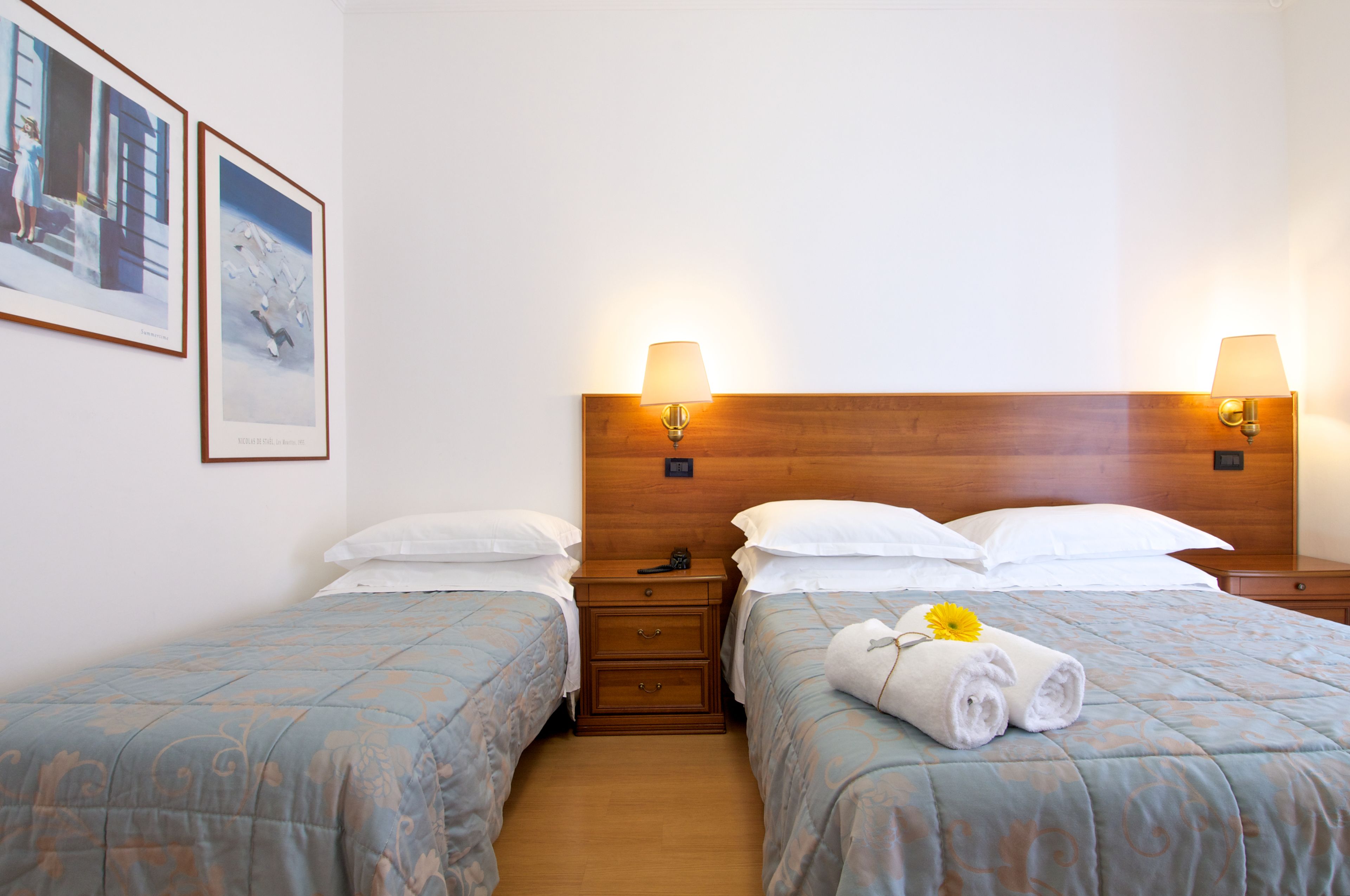 Camera d'albergo con due letti, poggiatesta in legno, lampade sui comodini e opere d'arte appese al muro.