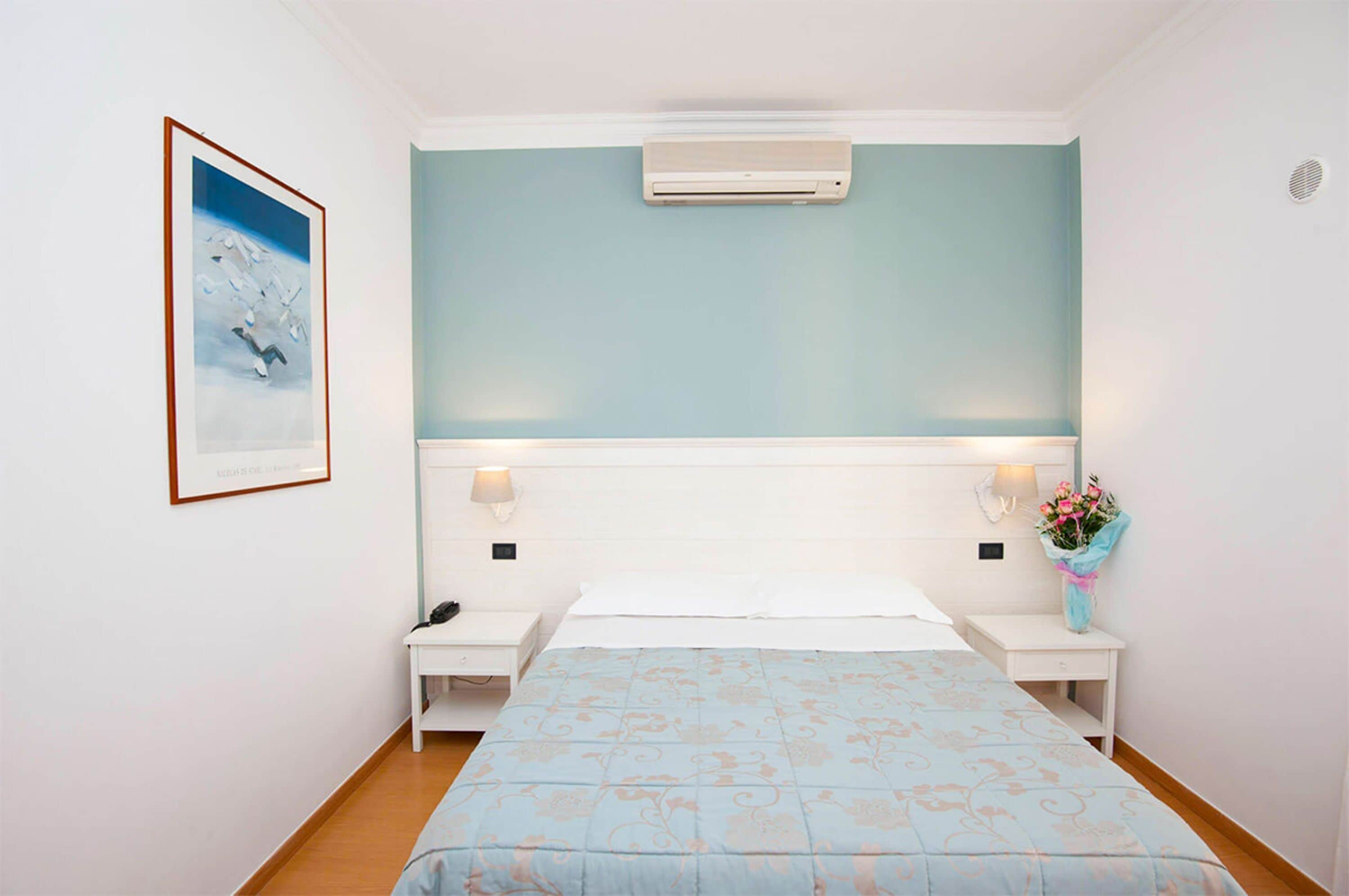 A bedroom with a bed, two bedside tables, a wall-mounted air conditioner, a framed picture, and a bouquet of flowers.