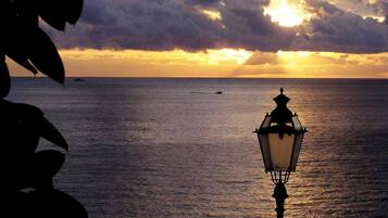 A silhouette of a person sitting on a bench with a lamp post in the foreground, overlooking a calm sea at sunset.