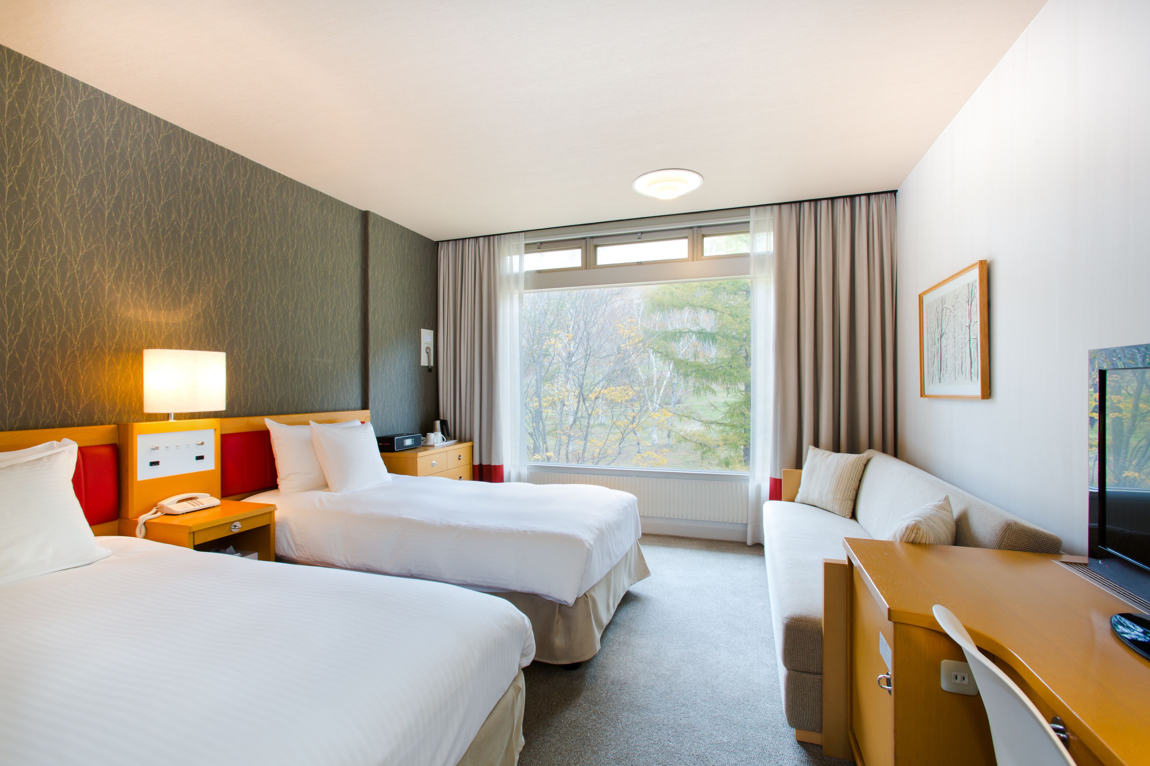 A hotel room with two beds, a desk, a TV, and a window with a view of trees.