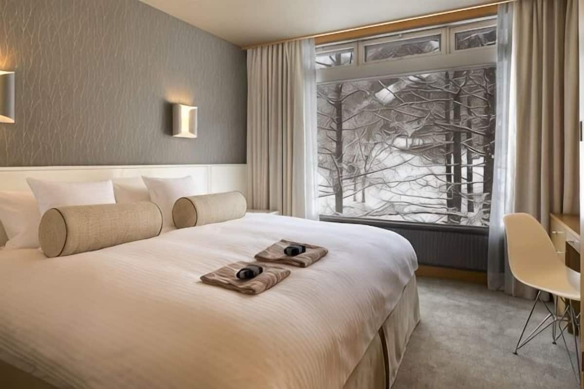 Ein Hotelzimmer mit einem großen Bett, einem Fenster mit Blick auf Bäume, einem Sessel und einem kleinen Tisch mit Tablett.