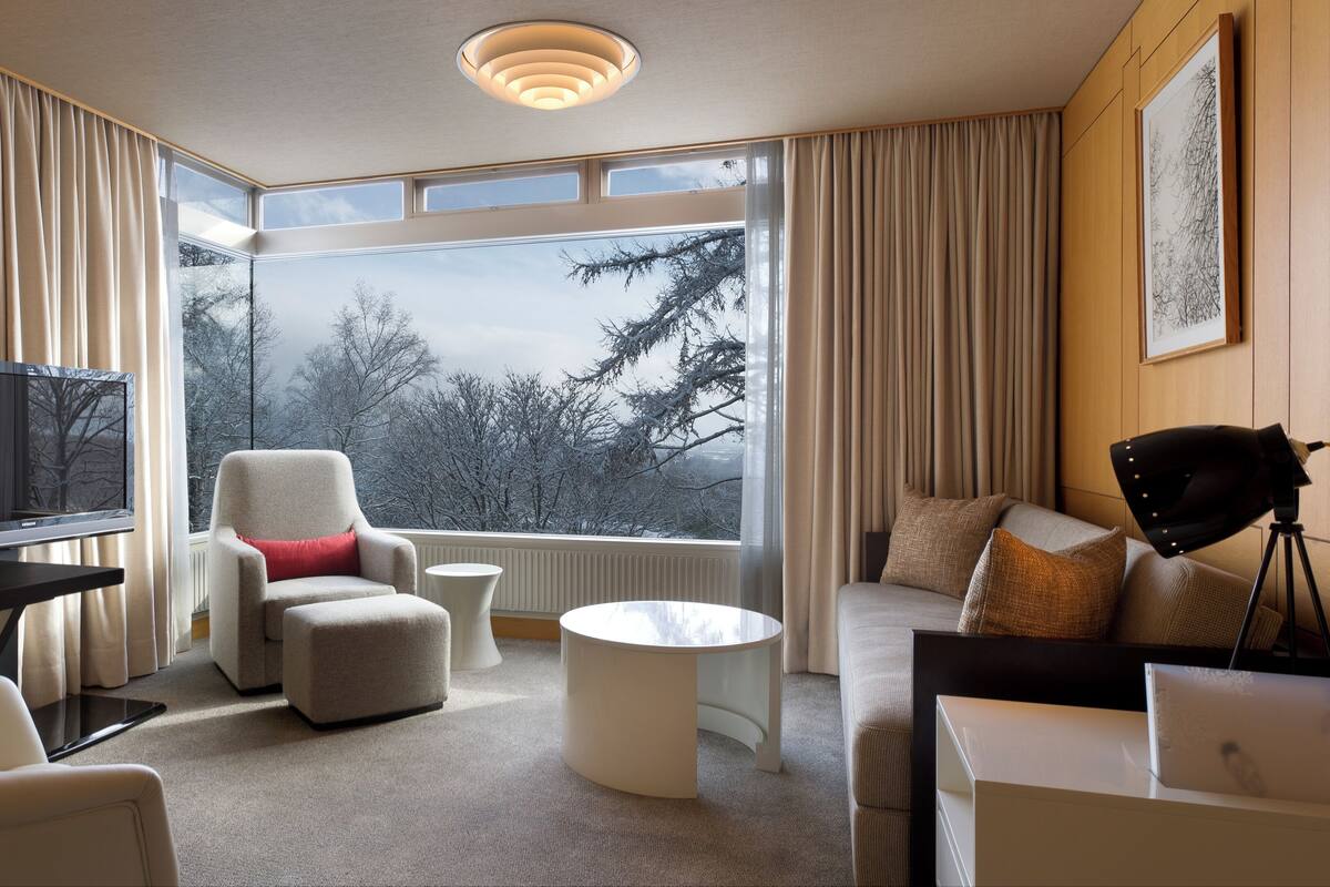 Ein modernes Hotelzimmer mit großem Fenster und Blick auf Bäume, einem Flachbildfernseher, einer gemütlichen Sitzecke und einem Schreibtisch mit Lampe.