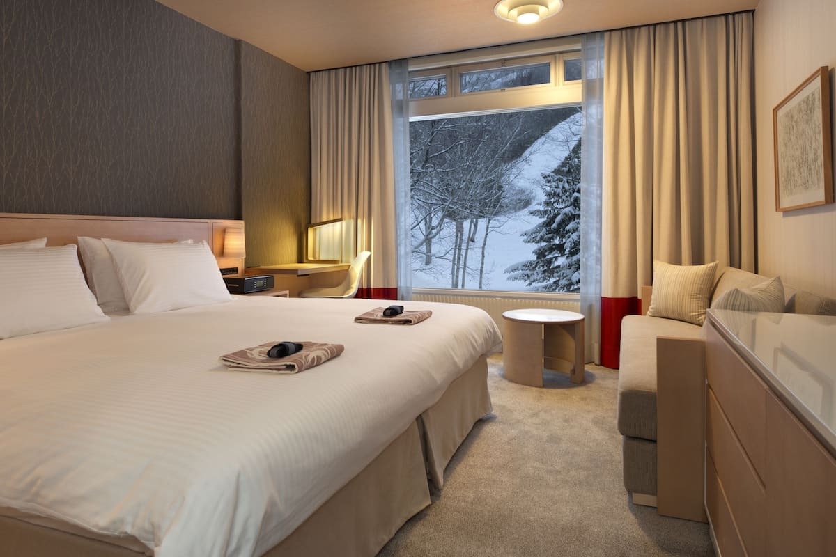 Ein Hotelzimmer mit einem großen Bett, einem Schreibtisch, einem Sessel, einem kleinen Tisch und einem Fenster mit Blick auf schneebedeckte Bäume.