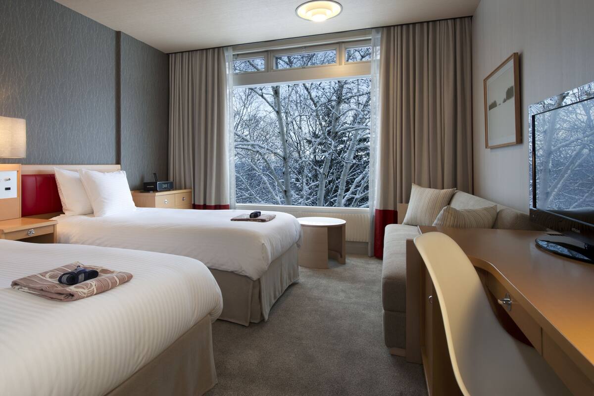 Ein Hotelzimmer mit zwei Betten, einem Schreibtisch, einem Sessel, einem Fernseher und einem Fenster mit Blick auf Bäume.