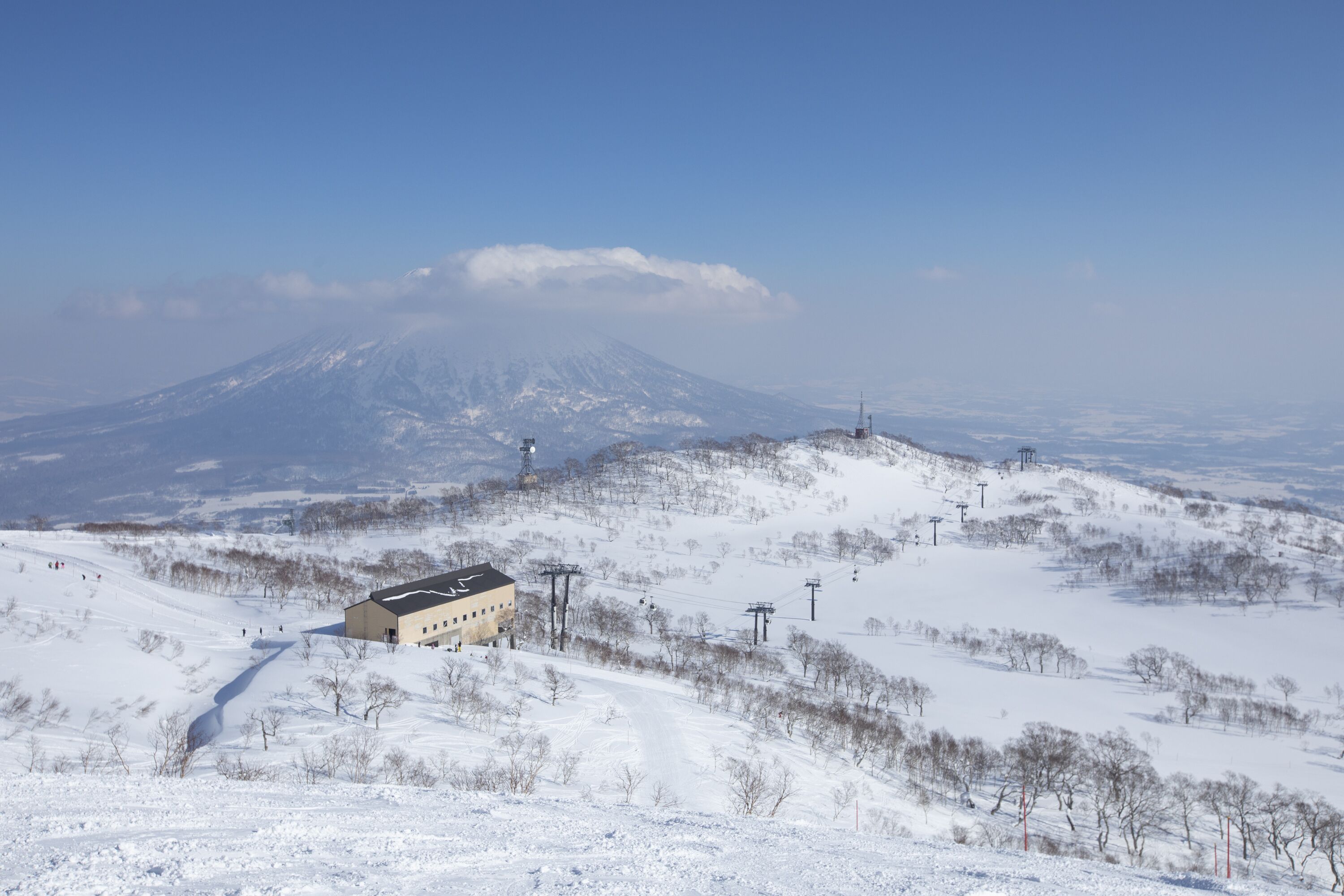 A snow-covered landscape with a mountain in the background, a building with a sloped roof, and ski lift cables.