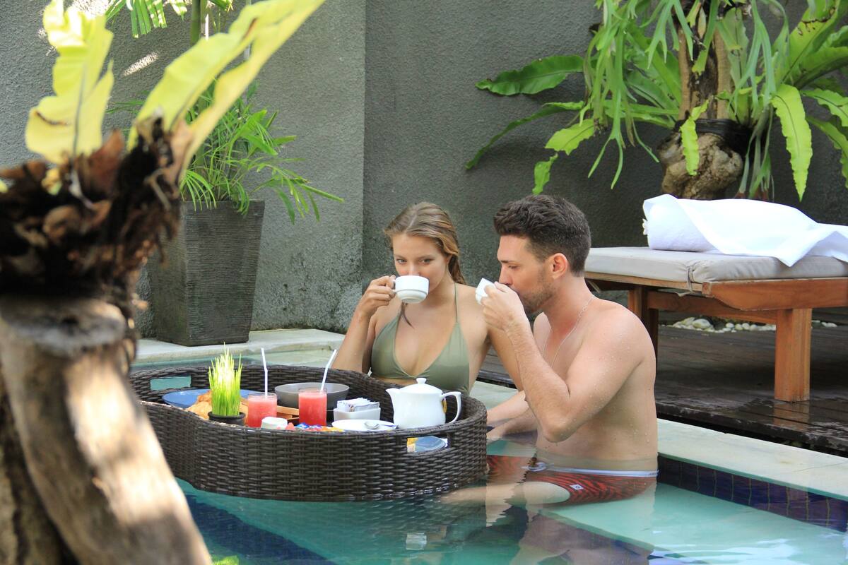 Um casal relaxando em uma piscina, com uma bandeja de vime contendo comida e bebidas.