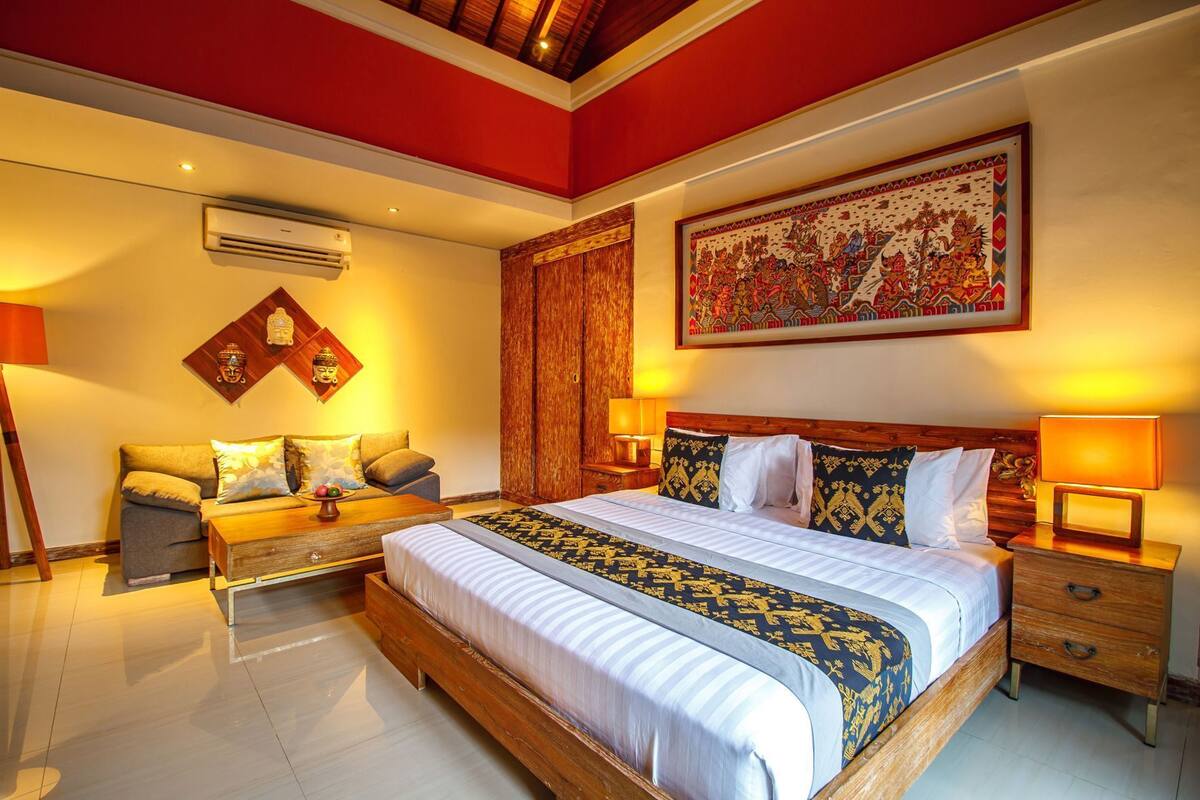 Quarto de hotel com uma cama grande, um sofá, uma cabeceira de madeira, ar condicionado fixado na parede e um quadro decorativo.