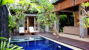 A swimming pool with a wooden deck, lounge chairs, and a stone wall. - The Wolas Villa & Spa (Seminyak)