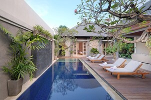 Piscina com espreguiçadeiras, rodeada por plantas em vasos e uma casa com telhado de azulejos.