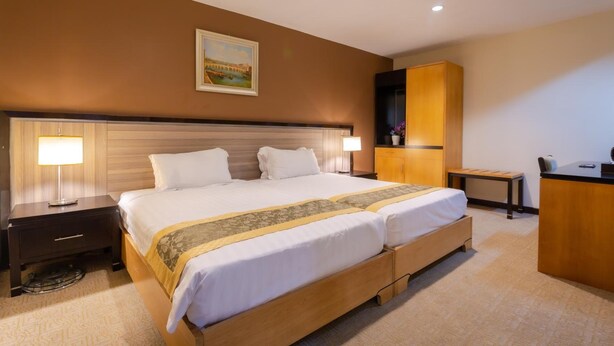 A hotel room with a large bed, two bedside tables with lamps, a wooden headboard, a desk with a chair, and a painting on the wall. - Hallmark View Hotel (Malacca City)