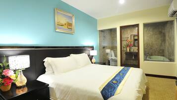 Quarto de hotel com uma cama grande, uma mesa de cabeceira com abajur, um quadro na parede e um banheiro visível através de uma porta aberta.
