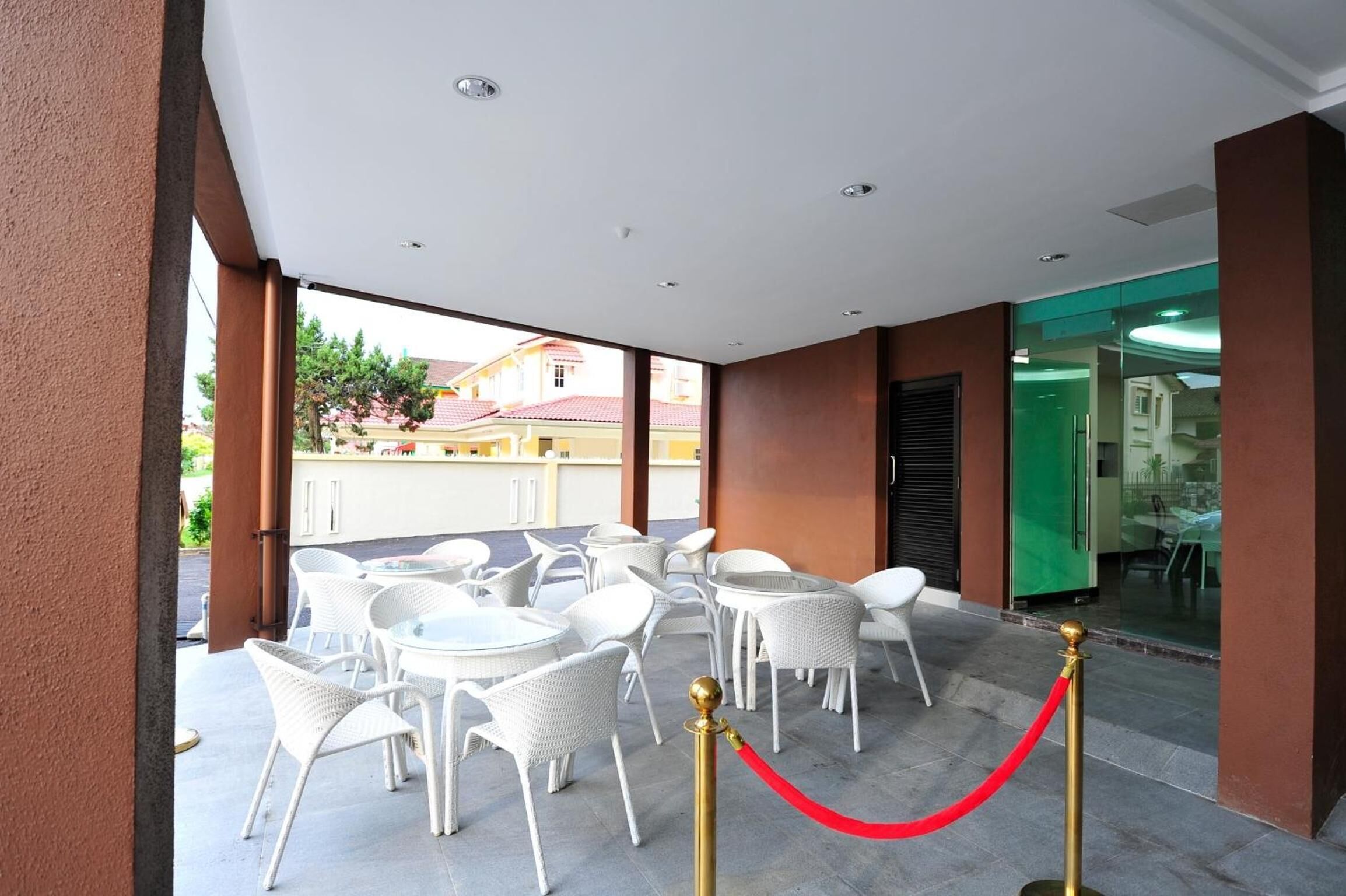 A covered outdoor seating area with white wicker chairs and tables, a red rope barrier, and a view of buildings in the background.