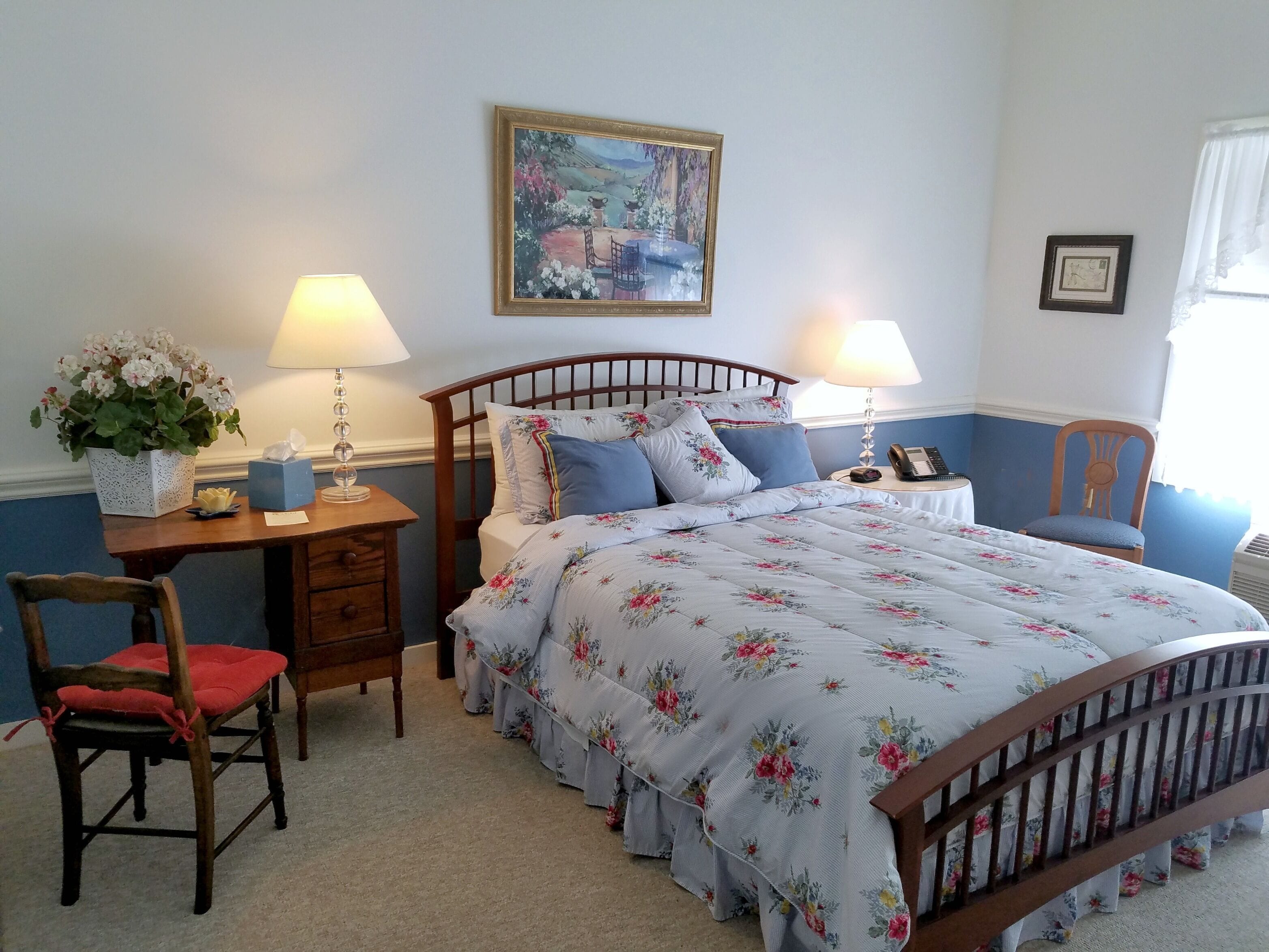 A neatly arranged bedroom with a bed, bedside tables, a chair, a desk, and a painting on the wall.