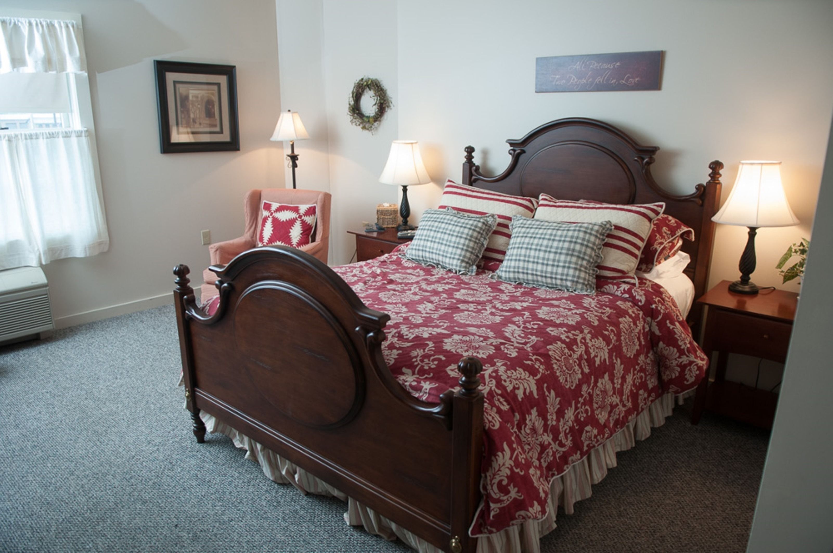 A neatly made bed with a wooden headboard, two bedside tables with lamps, a framed picture on the wall, and a window with curtains.