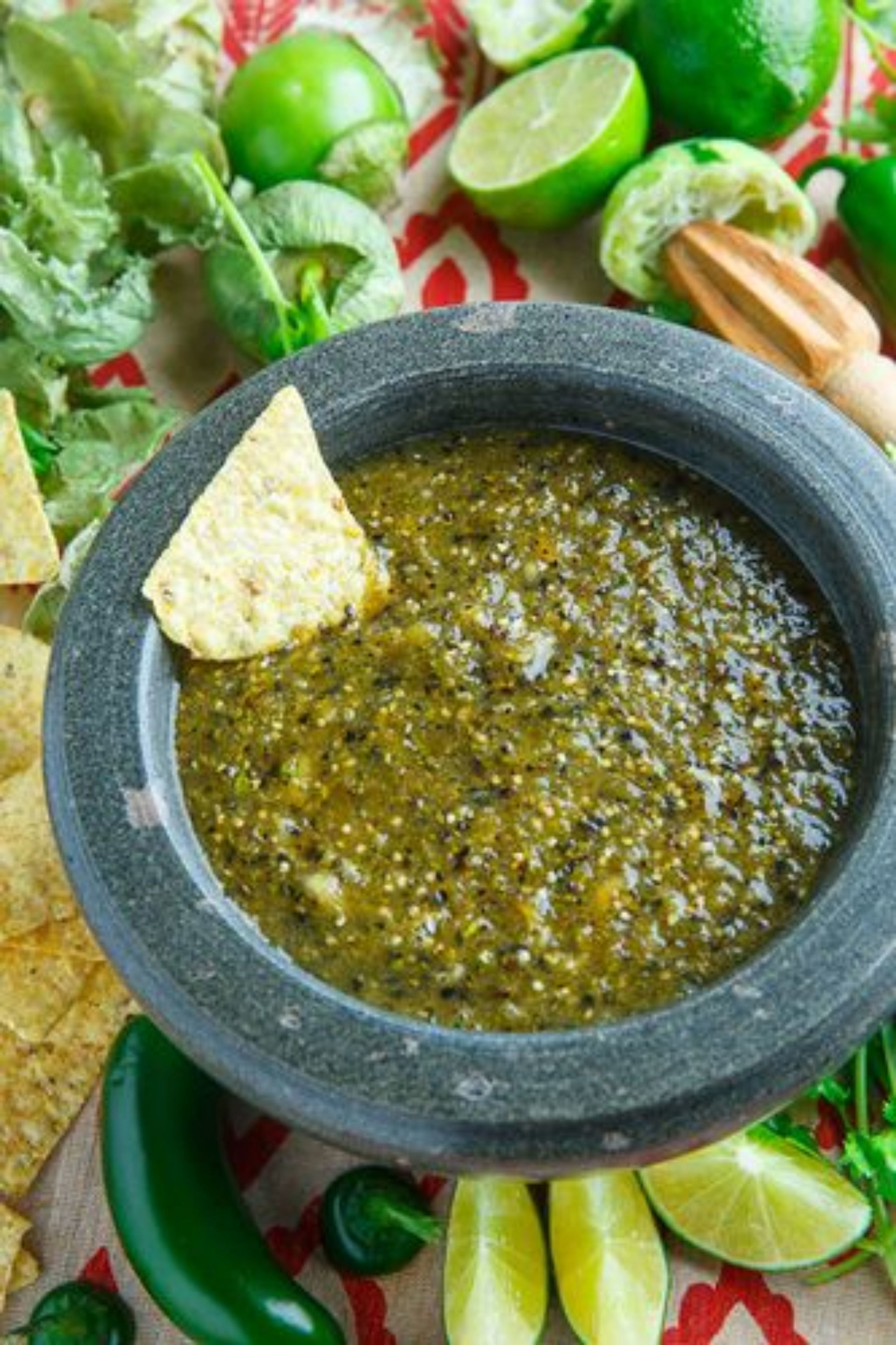 A bowl of green sauce with a tortilla chip, surrounded by lime, chili peppers, and lettuce.