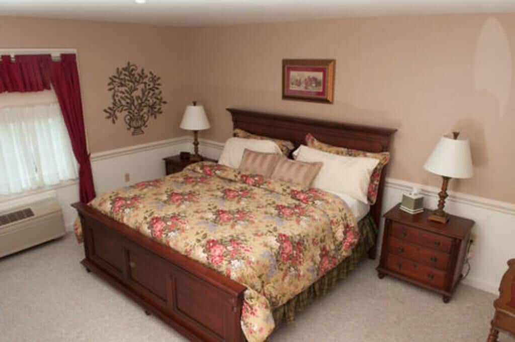 A bedroom with a wooden bed, floral bedding, two bedside tables with lamps, a wall-mounted picture, and a window with curtains.