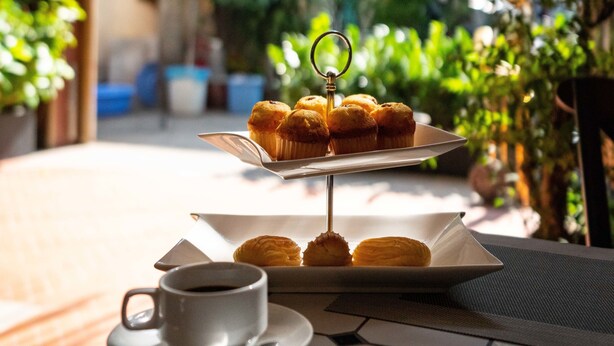 Una mesa con una taza blanca y pasteles, en un espacio exterior soleado con plantas y un estante metálico al fondo.