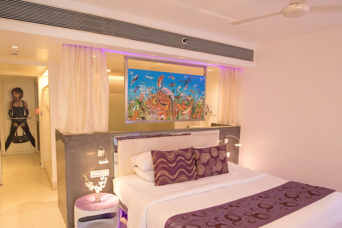 Quarto de hotel moderno com uma cama grande, um quadro acima da cama, um ventilador de teto e um banheiro visível ao fundo.