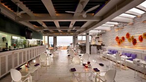 Un restaurante moderno con vistas a la playa, que cuenta con mobiliario blanco y una zona de bar.