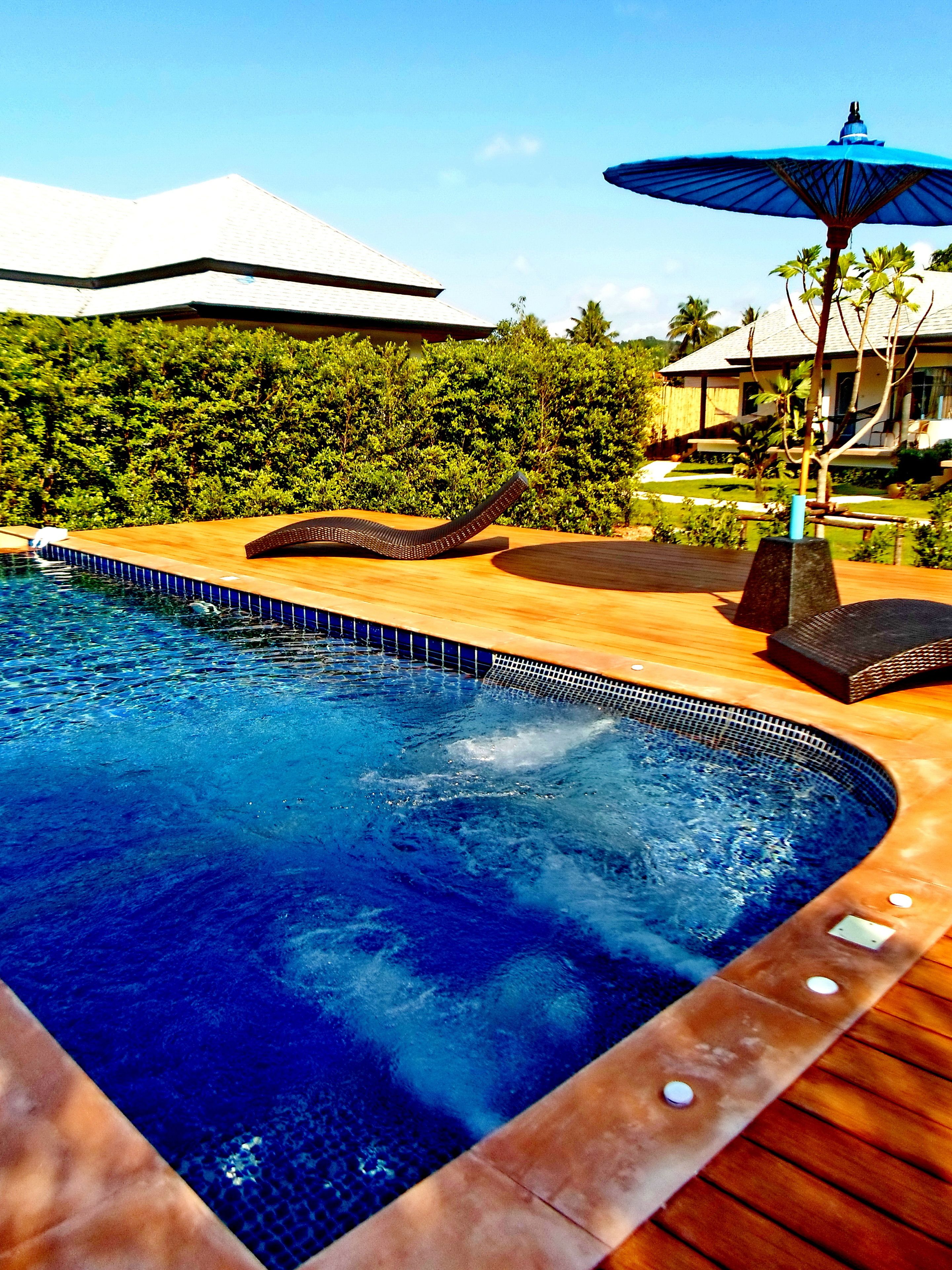 Una piscina con muelle de madera, sillas de descanso y una sombrilla azul.