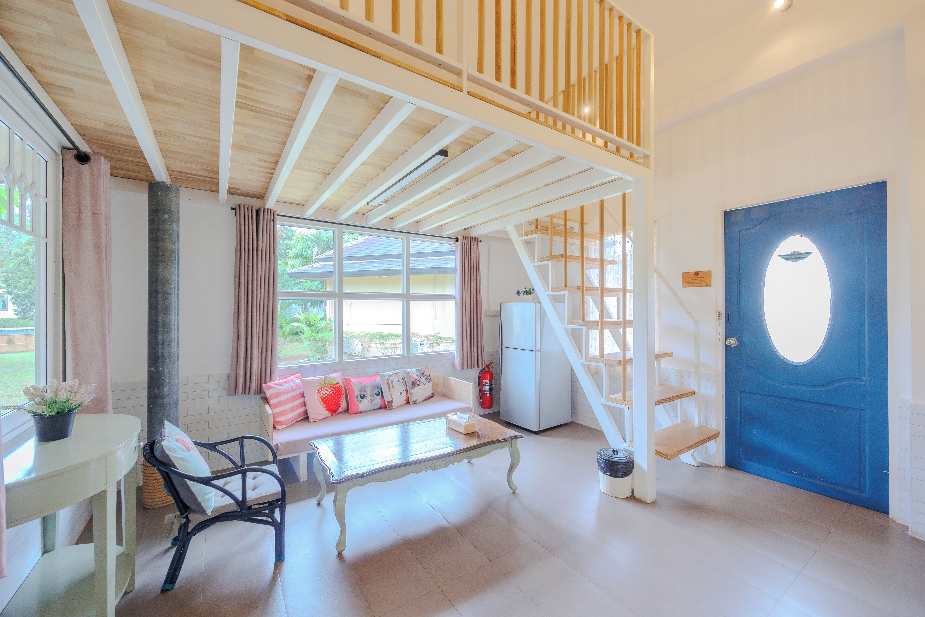 Una habitación amplia con techo de madera, una puerta azul, una escalera, un rincón de descanso con cojines y una mesa.