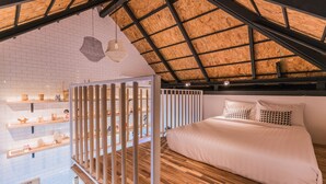 Een slaapkamer met een schuine kap, houten vloeren, een bed met wit beddengoed en een witte wand met planken waarop diverse spullen staan.