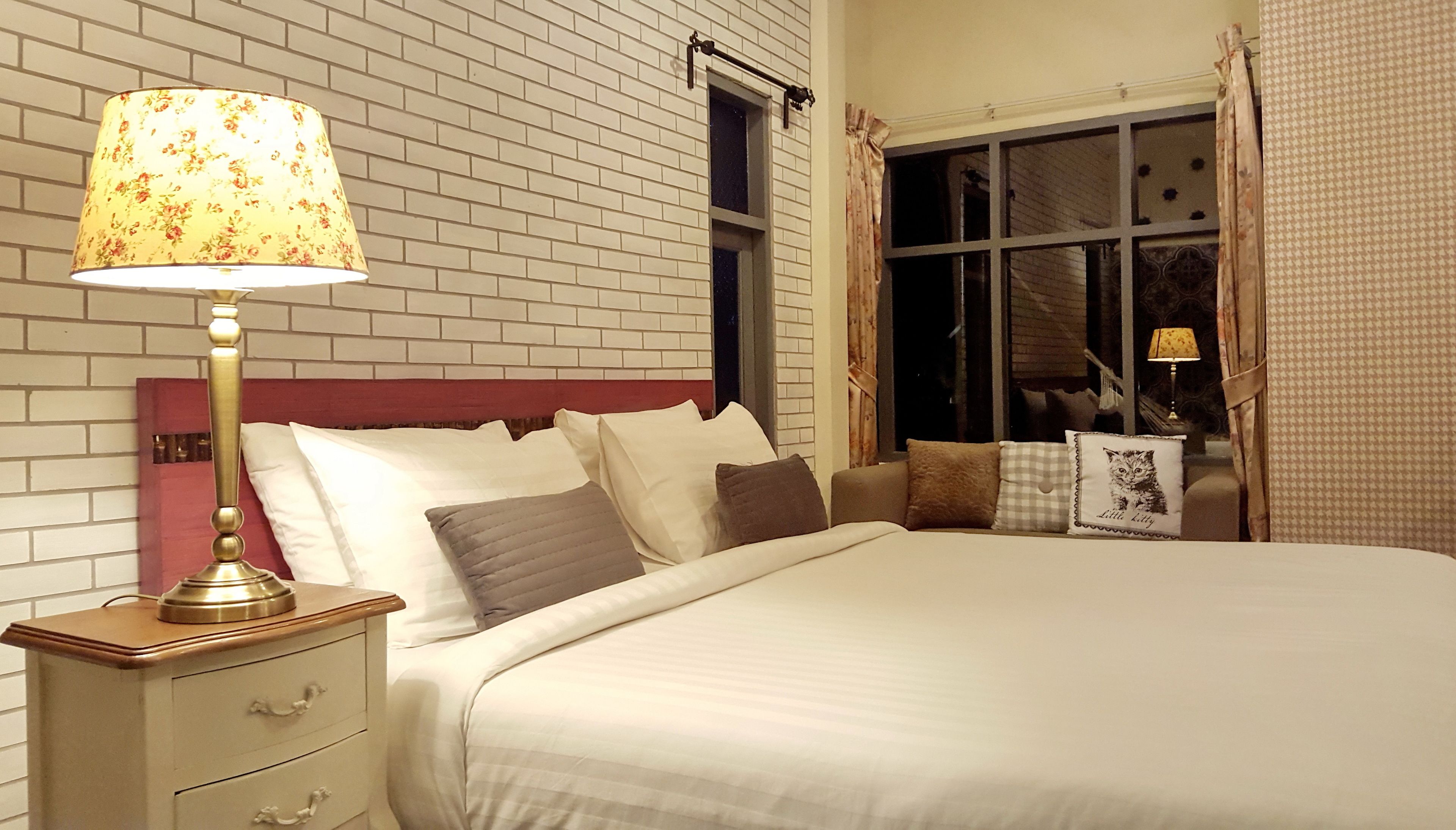 A neatly made bed with white linens, a wooden nightstand with a lamp, and a window with curtains.