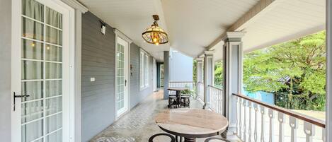 A covered porch with a round table and four stools, a hanging light fixture, and a view of greenery.