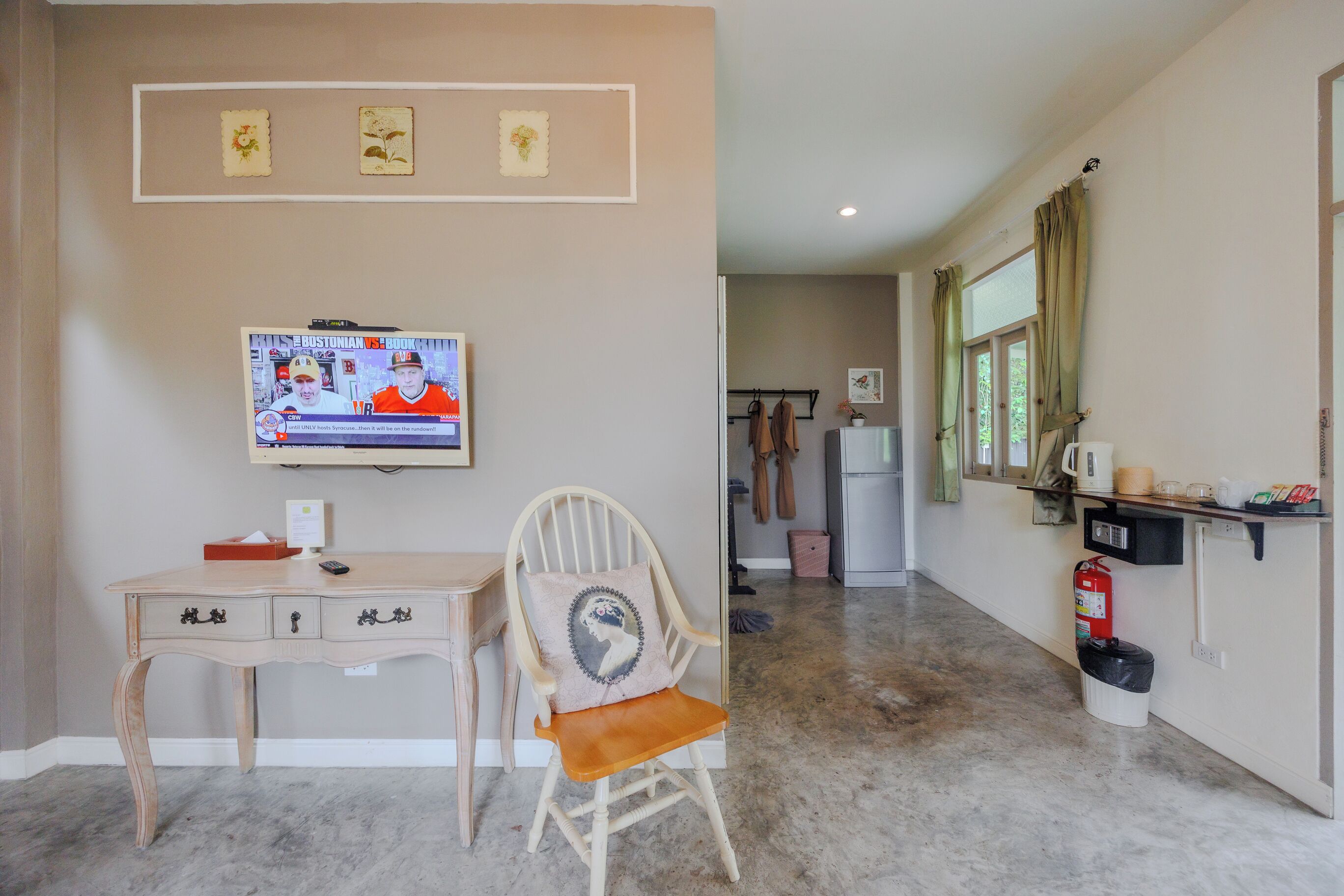 A room with a wooden desk, a cushioned chair, a television, and a hallway leading to a kitchen area.