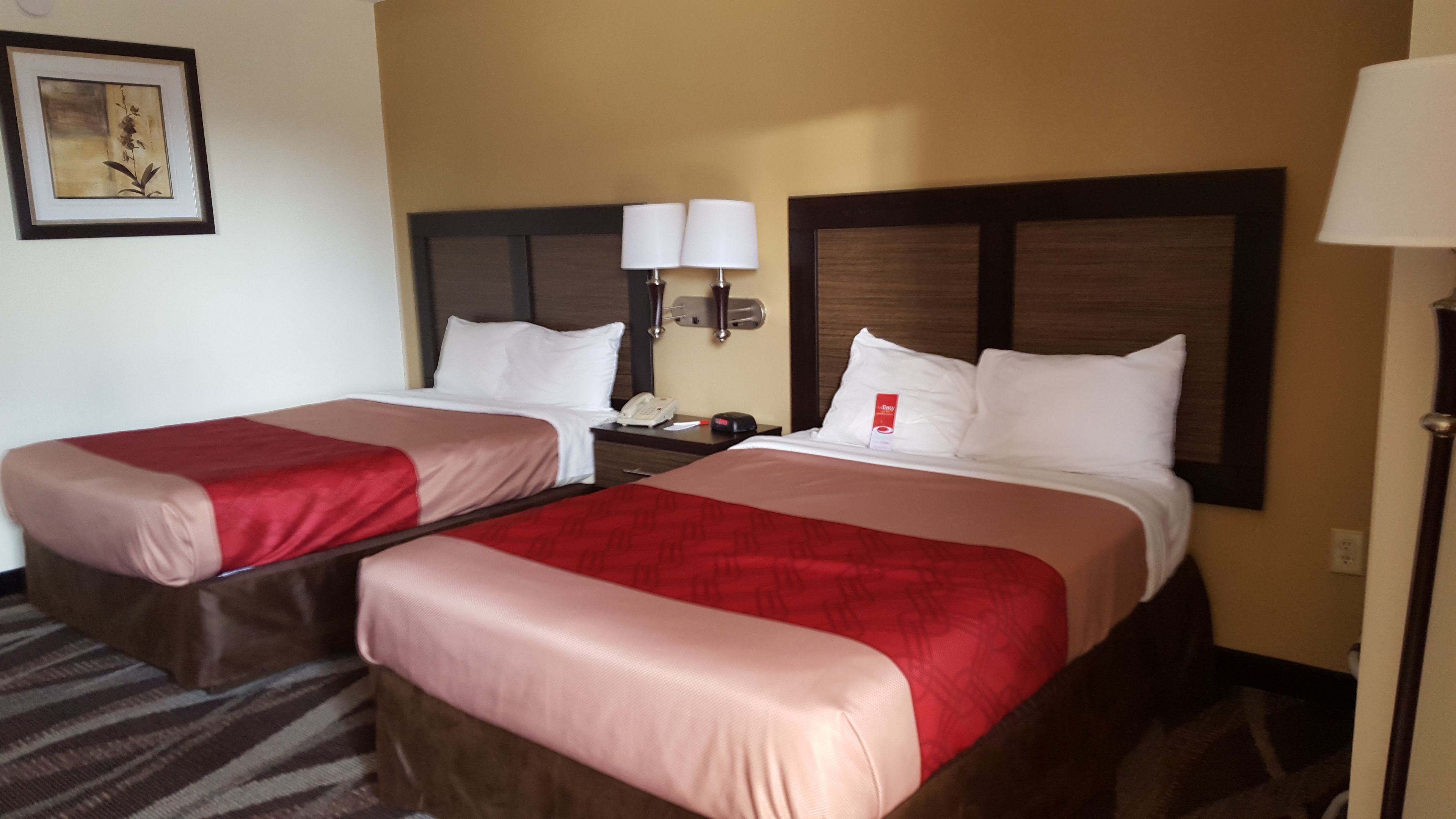 A hotel room with two beds, each with a red and beige bedspread, white pillows, and a nightstand with a lamp and a phone.
