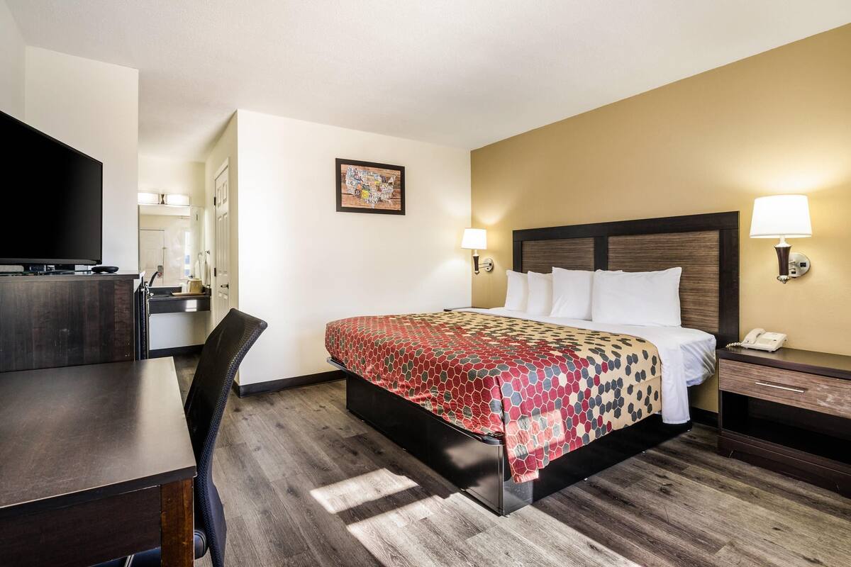 A hotel room with a bed, bedside table, lamp, and a TV on a desk.