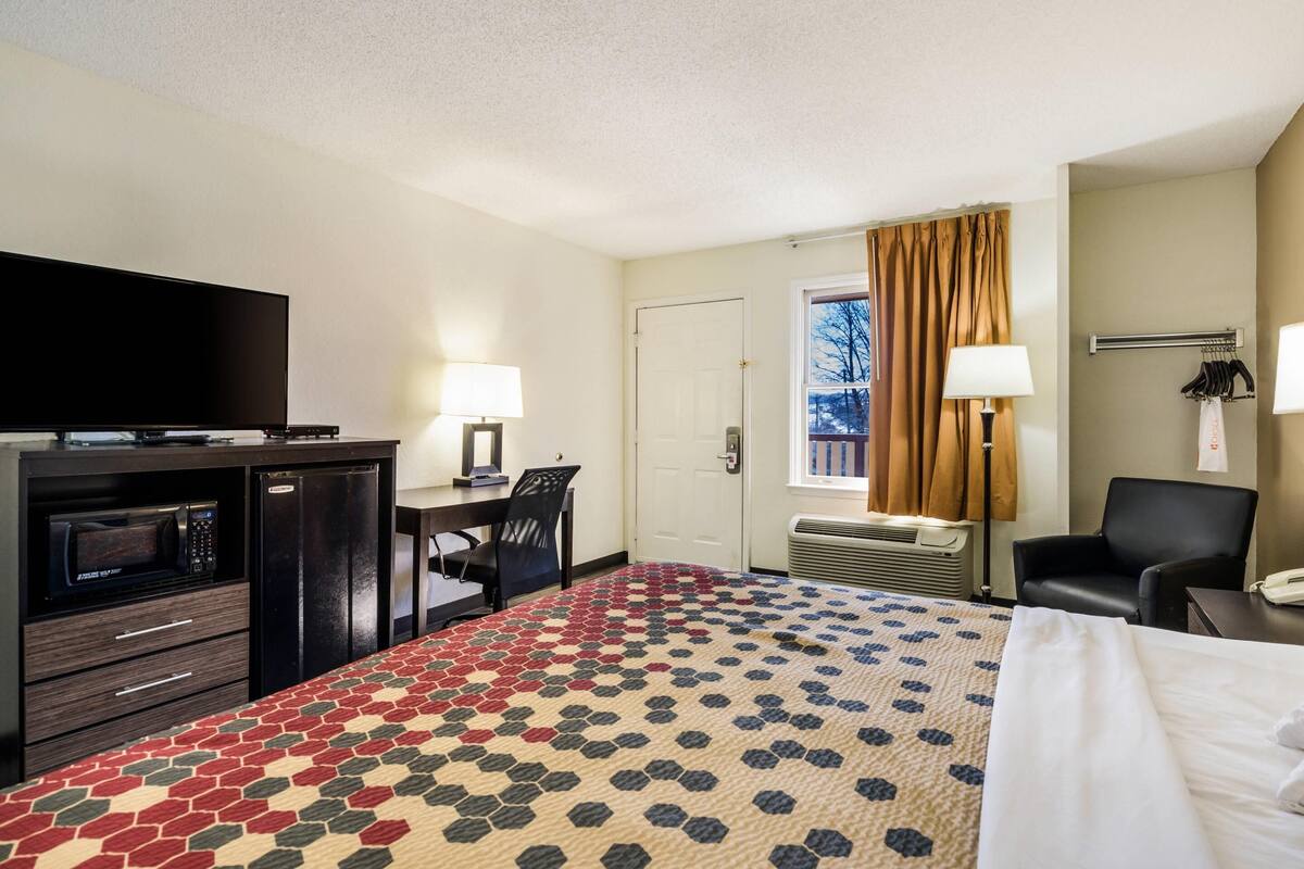 A hotel room with a bed, a desk with a chair, a television, a microwave, and a window with curtains.